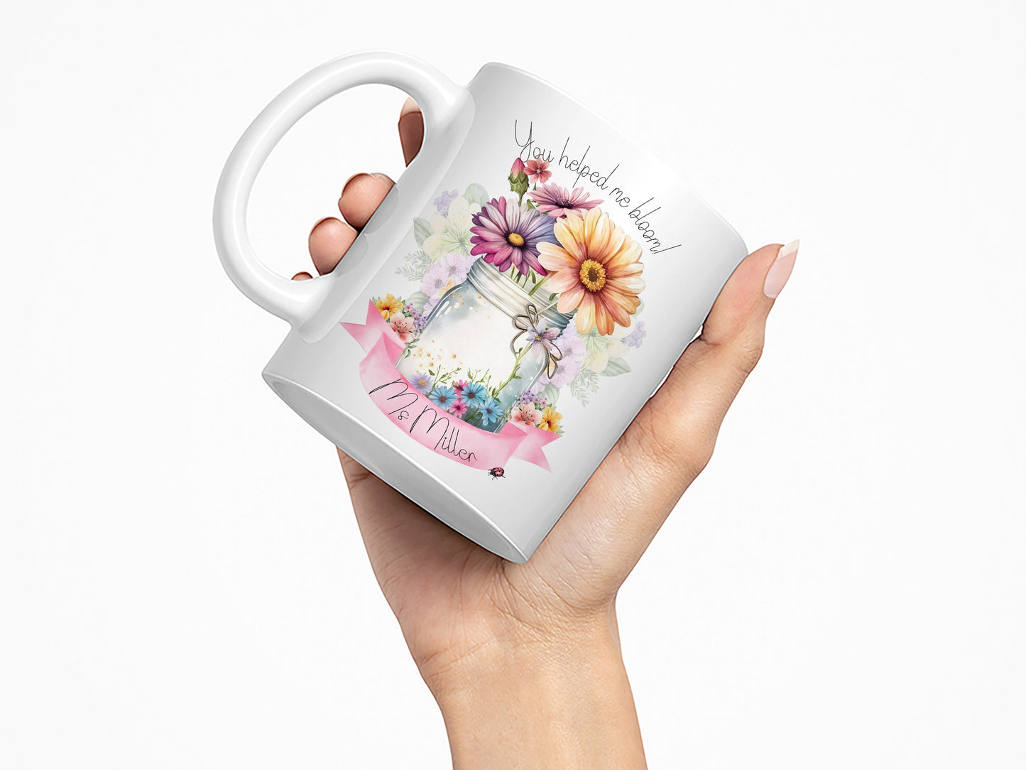 White Ceramic mug. A jar vase holding a variety of summer flowers and a pink banner across bottom Text reads You helped me bloom! arched around top of flowers and Name is written within the banner