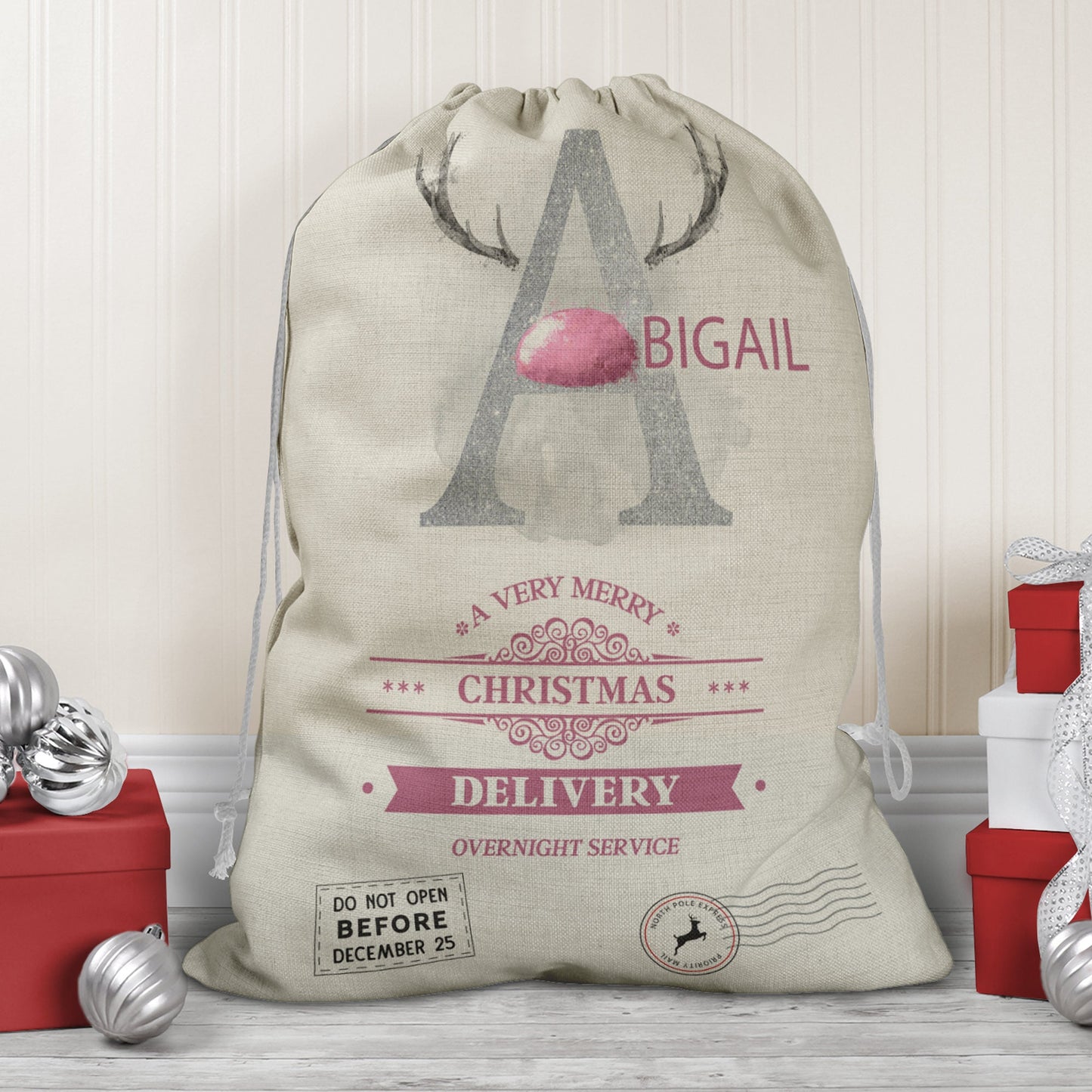 Large natural looking Linen Sack with printed Reindeer antler and Silver Initial letter with pink coloured nose & text Name is written in pink above the words A very merry Christmas Delivery Overnight service and two North pole style delivery stamps