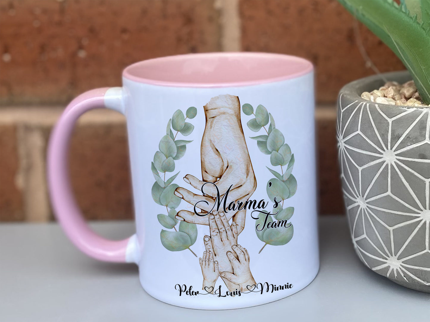 Ceramic mug with pastel pink handle and rim. Printed with watercolour style artwork of adult female hand and 3 childrens hands