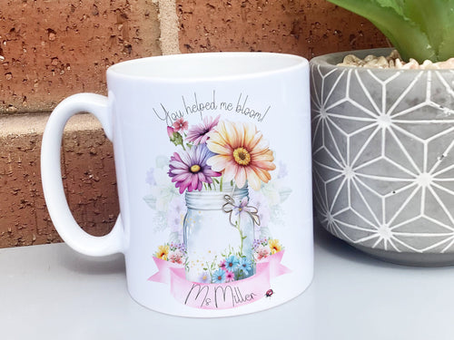 White Ceramic mug. A jar vase holding a variety of summer flowers and a pink banner across bottom Text reads You helped me bloom! arched around top of flowers and Name is written within the banner