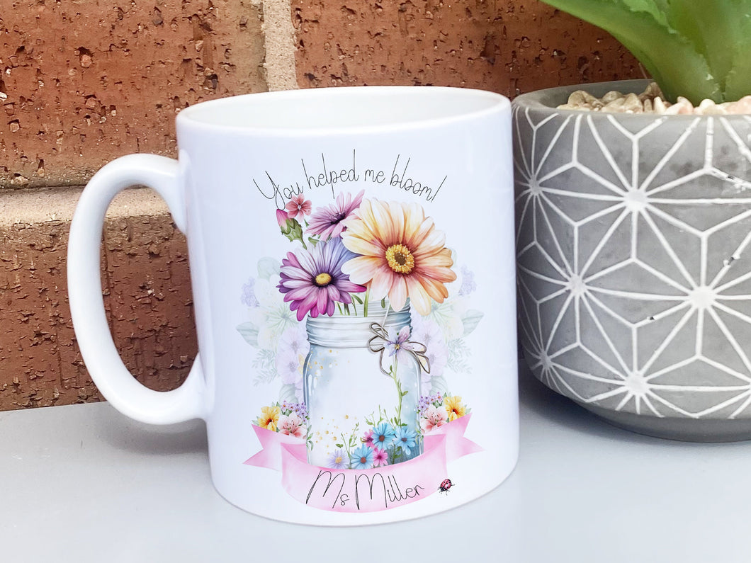 White Ceramic mug. A jar vase holding a variety of summer flowers and a pink banner across bottom Text reads You helped me bloom! arched around top of flowers and Name is written within the banner
