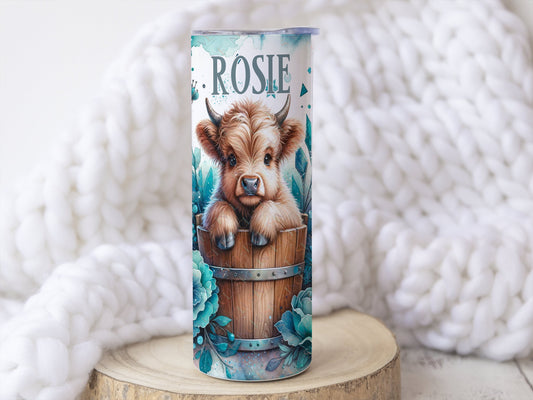 20oz white stainless teel tumbler with a realistic baby highland cow sitting in a wooden barrel image  blue flowers complete the design which wraps around the cup. Matching text for name can be added to bottom of tumbler