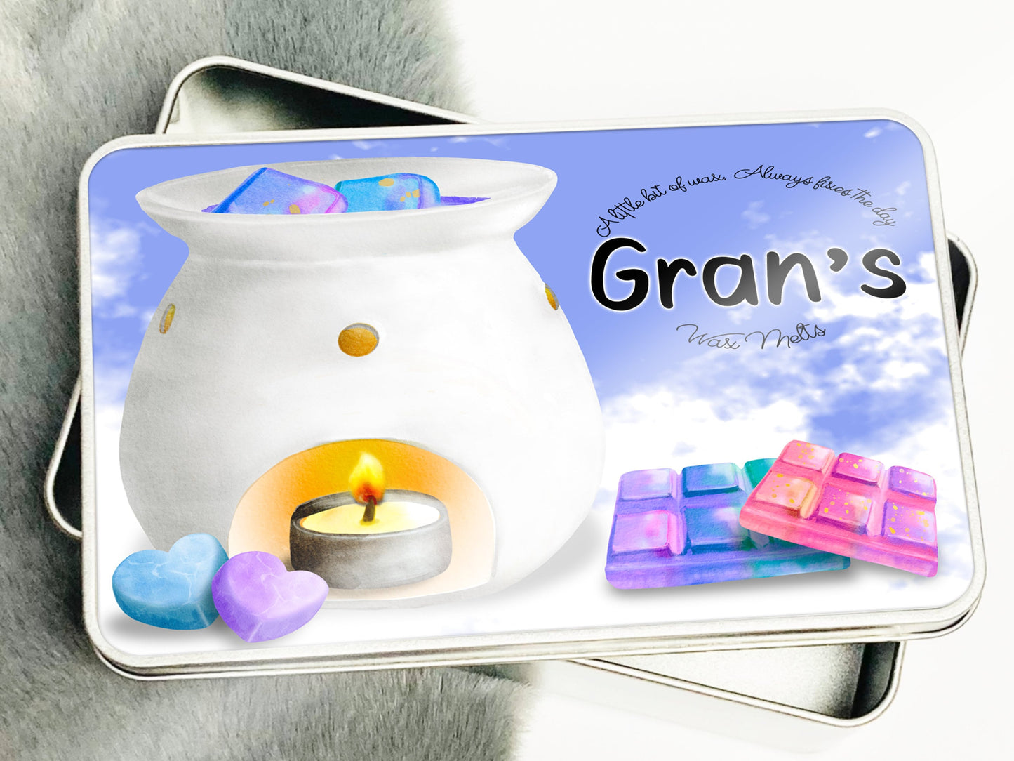 Image shows a Silver metal Rectangular Personalised Wax melt storage tin.sitting on a grey and white faux fur rug.  Text reads A little bit of wax Always fixes the day. Grans Wax melts