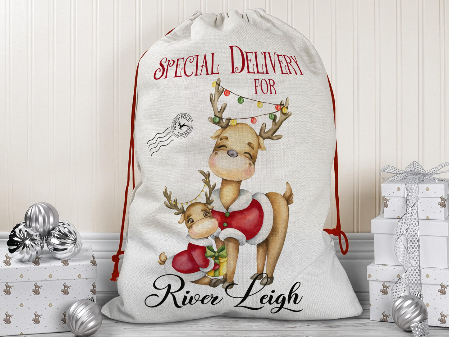 Personalized Christmas Santa sack featuring a cute parent-and-child reindeer illustration with festive lights, "Special Delivery for" text, and custom name "River Leigh" printed at the bottom. The sack has a red drawstring closure and is styled as a gift bag placed among silver ornaments and holiday-wrapped presents.