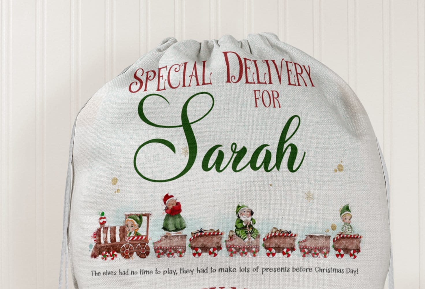 Close up of the top half of Large linen sack with red drawstring and red and green print

Text reads 
Special Delivery for 
NAME

Underneath sits a festive train carrying elves in green and red with candy cane coloured wheels