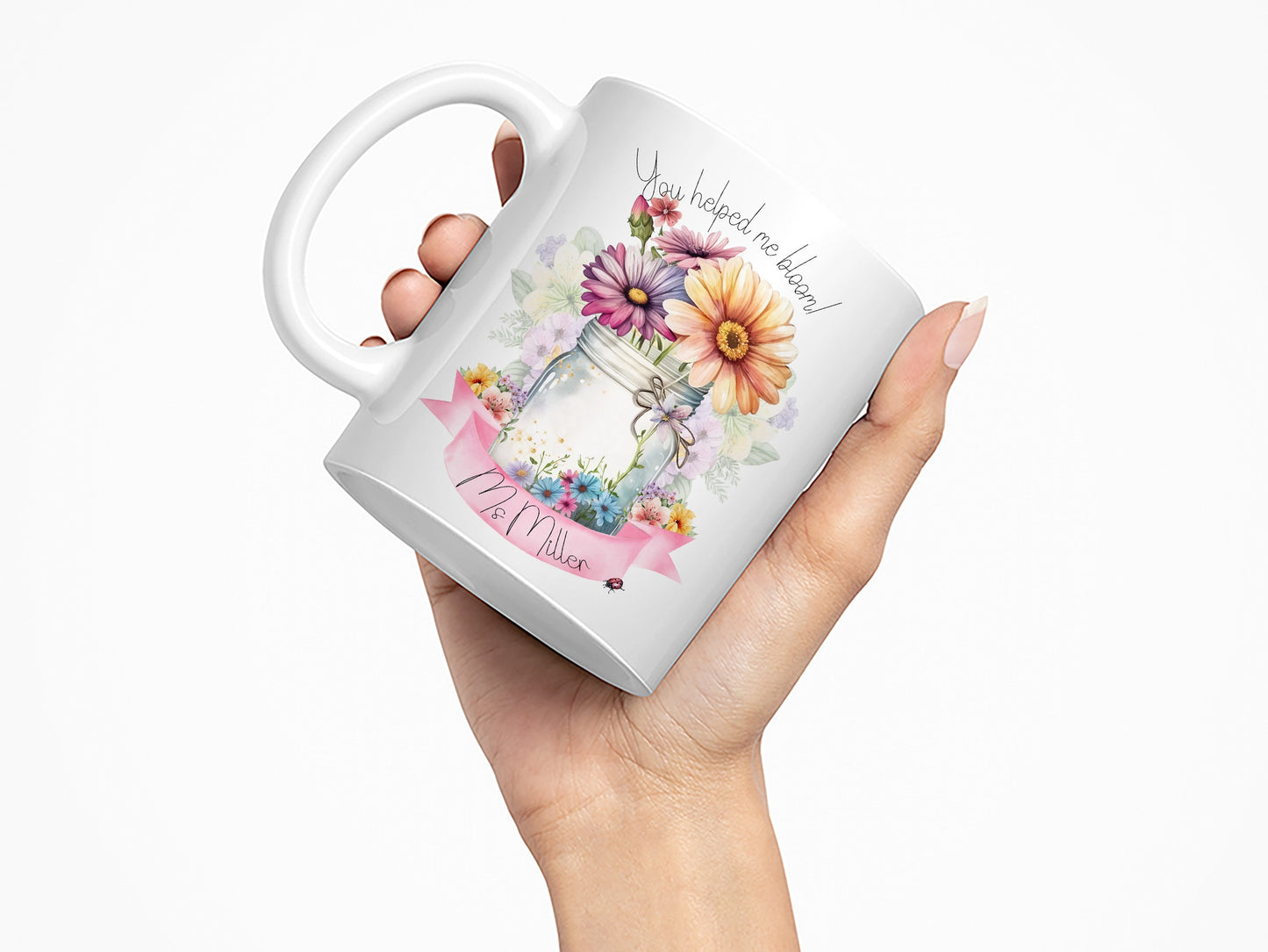White Ceramic mug. A jar vase holding a variety of summer flowers and a pink banner across bottom Text reads You helped me bloom! arched around top of flowers and Name is written within the banner