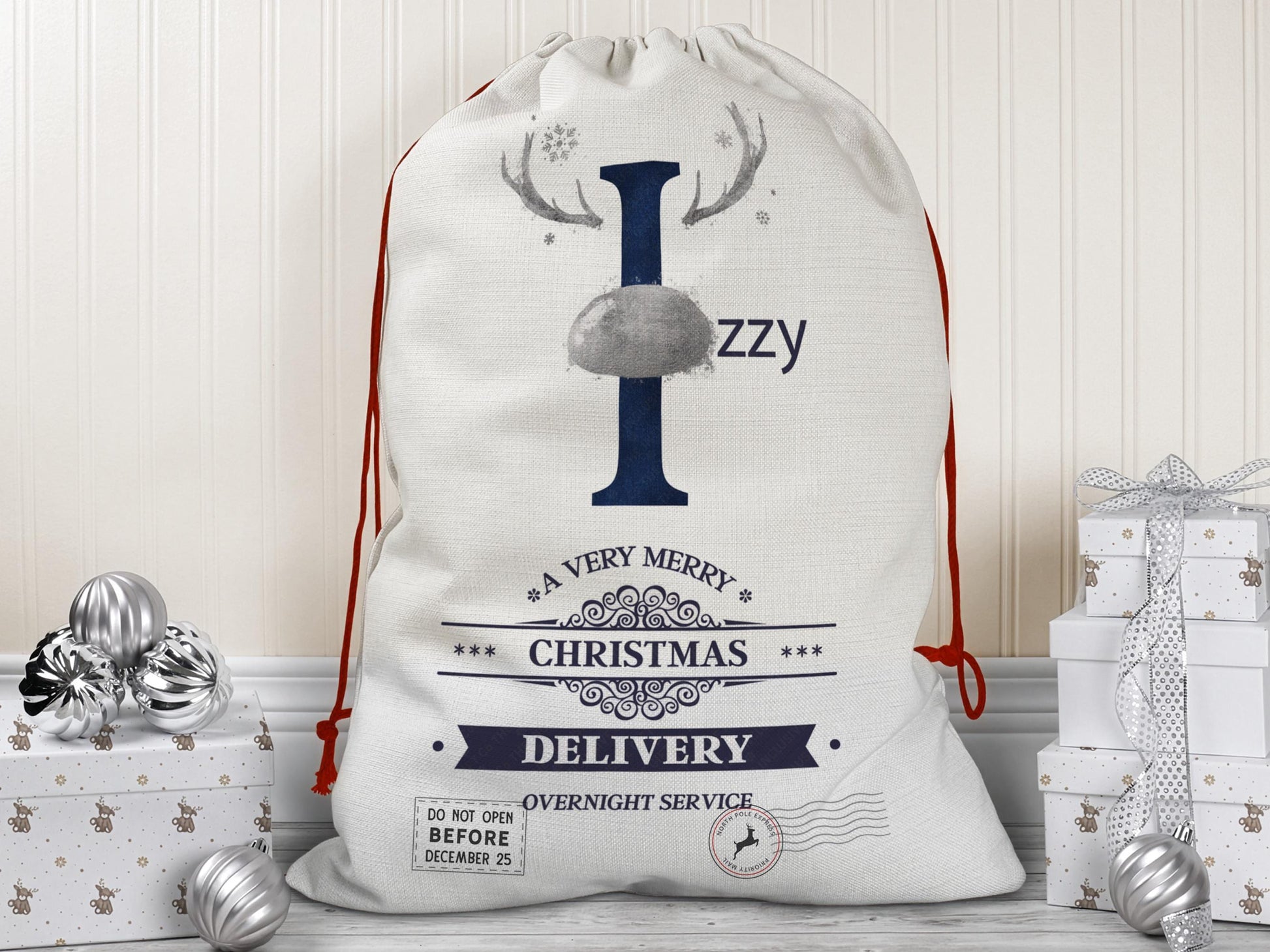 A large beige Christmas gift sack with red drawstrings stands upright on a wood floor against a cream-colored wall. The sack features a large Navy letter “I” decorated with Silver reindeer antlers and a matching Silver nose