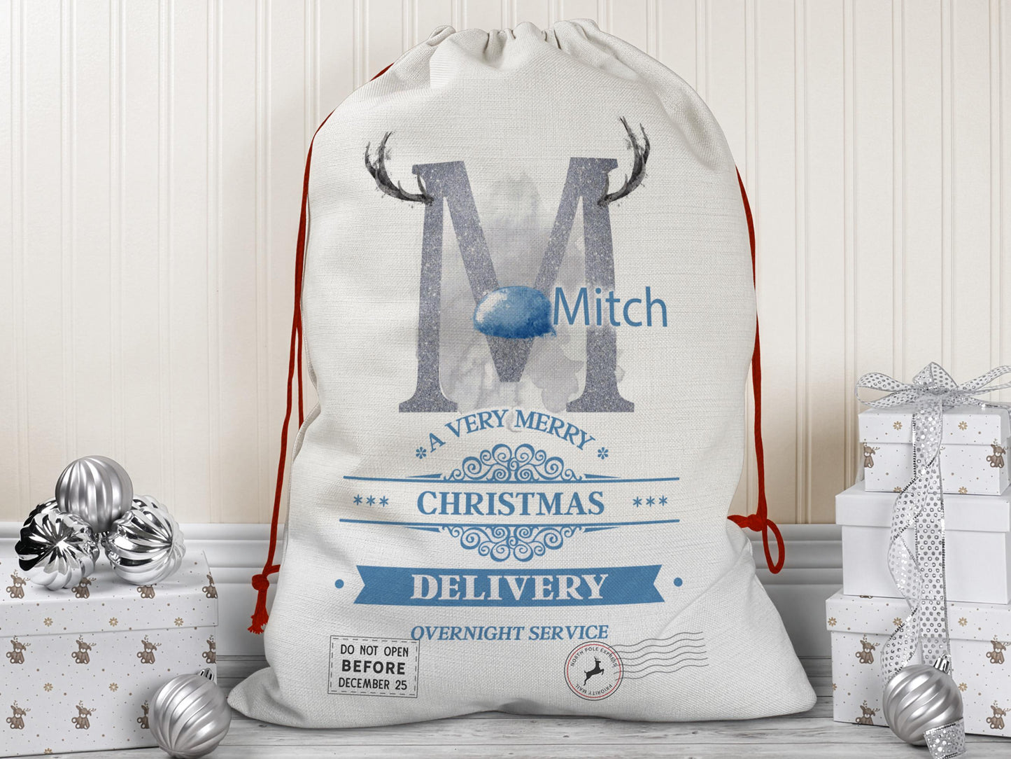 A large beige Christmas gift sack with red drawstrings stands upright on a wood floor against a cream-colored wall. The sack features a large Baby Blue letter “M” decorated with reindeer antlers and a matching Blue nose.