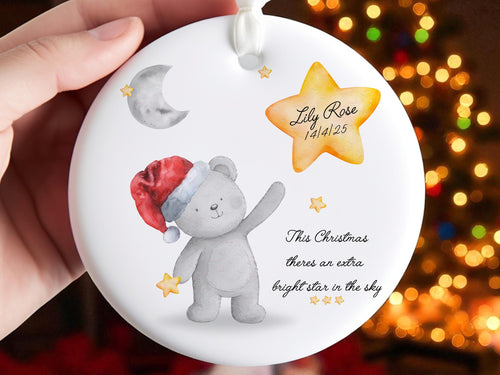 Bright white flat circle ceramic ornament with white satin ribbon. Printed with a gentle comforting remberance design featuring a soft grey bear wearing a santa hat and clutching a gold star standing under a greay crescent moon pointing up to a bright yellow Gold shining star featuring baby or childs name and relevant dates. Black text sits below the star that reads "This Christmas theres an extra bright star in the sky."