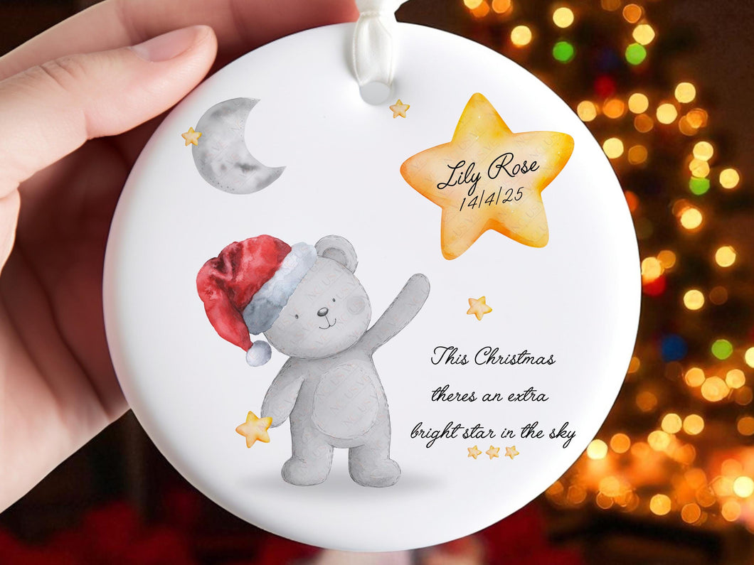 Bright white flat circle ceramic ornament with white satin ribbon. Printed with a gentle comforting remberance design featuring a soft grey bear wearing a santa hat and clutching a gold star standing under a greay crescent moon pointing up to a bright yellow Gold shining star featuring baby or childs name and relevant dates. Black text sits below the star that reads "This Christmas theres an extra bright star in the sky."