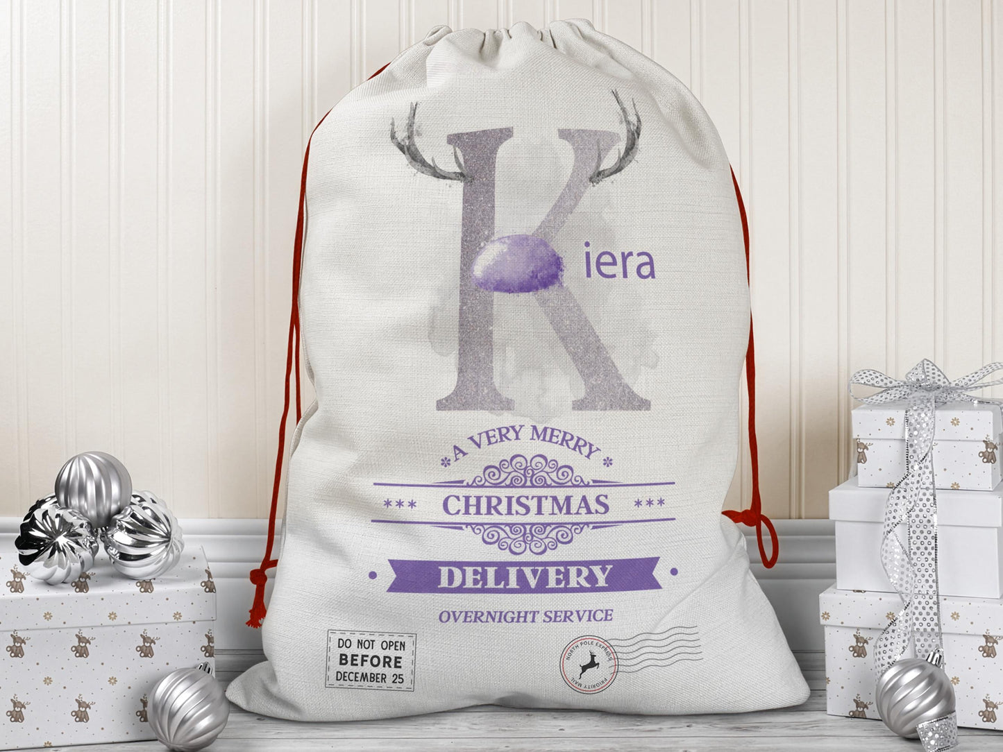 A large beige Christmas gift sack with red drawstrings stands upright on a wood floor against a cream-colored wall. The sack features a large Lilac letter “K” decorated with reindeer antlers and a matching Lilac nose