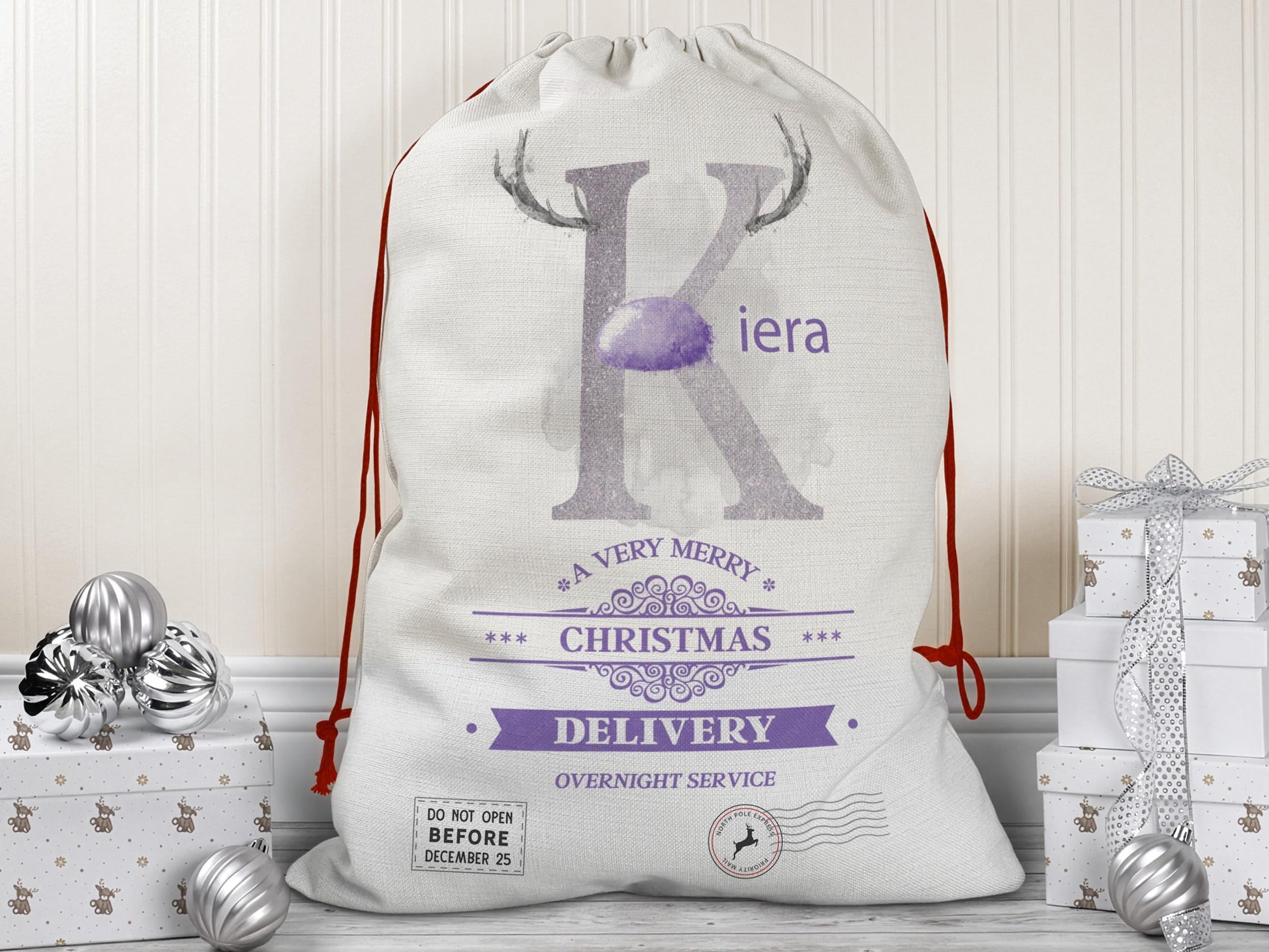A large beige Christmas gift sack with red drawstrings stands upright on a wood floor against a cream-colored wall. The sack features a large Lilac letter “K” decorated with reindeer antlers and a matching Lilac nose