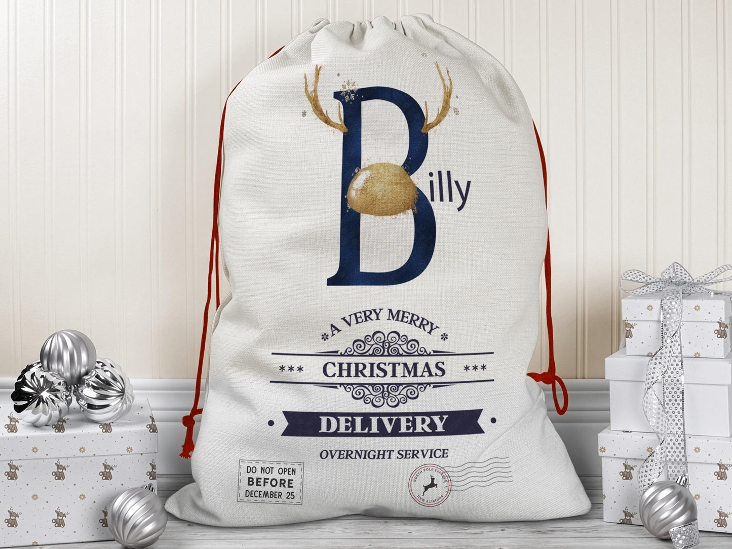 A large beige Christmas gift sack with red drawstrings stands upright on a wood floor against a cream-colored wall. The sack features a large Blush Navy letter “B” decorated with Gold reindeer antlers and a matching Gold nose