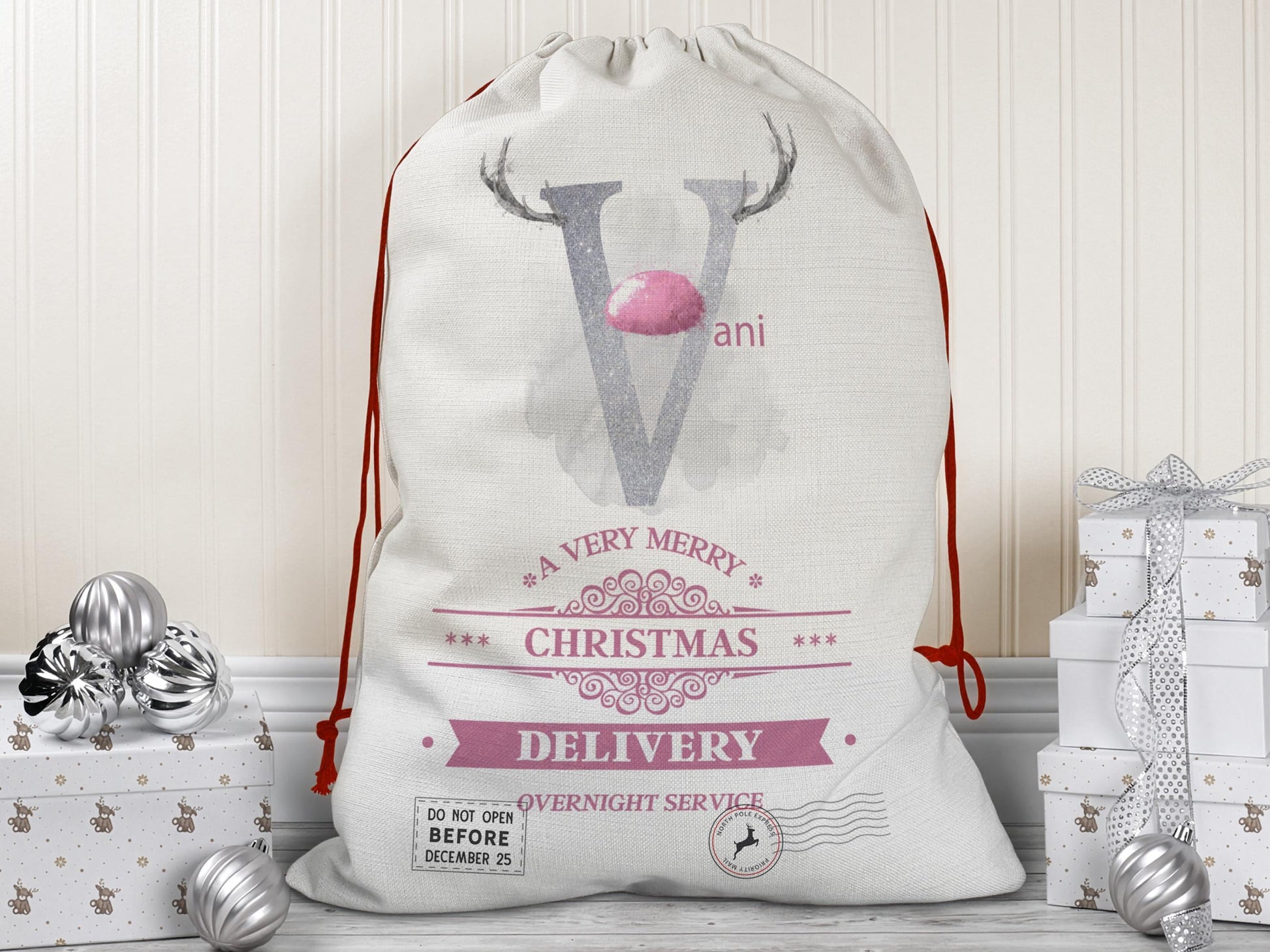 A large beige Christmas gift sack with red drawstrings stands upright on a wood floor against a cream-colored wall. The sack features a large Blush Pink letter “V” decorated with reindeer antlers and a Pink nose, along with the name “Vani” beside it. Below the letter is festive pink typography reading “A Very Merry Christmas Delivery – Overnight Service.” Silver ornaments and white gift boxes with subtle holiday patterns are placed on the floor around the sack. 5 other colours available