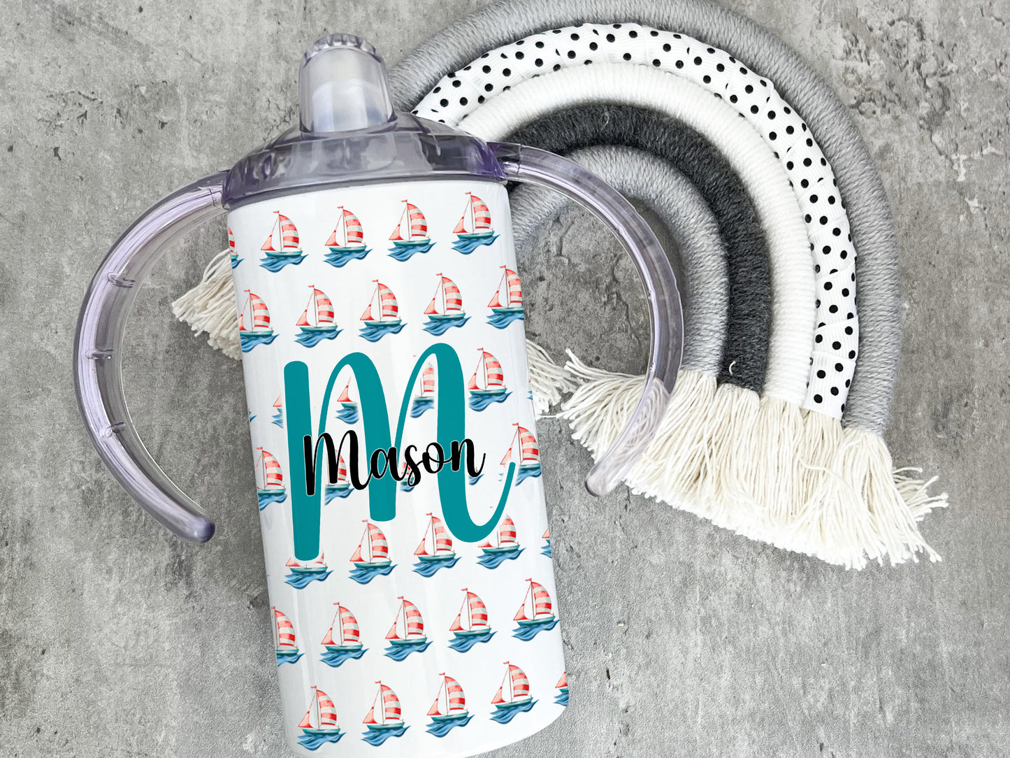 White Adult sippy cup with sailing boat design and inital and name. Image shows single cup on laying on grey rainbow prop. Design shows sailing boat pattern and Large Initial with name across the middle.