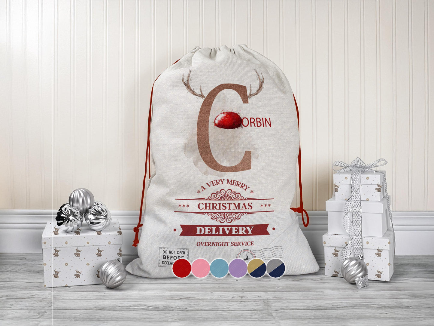 A large beige Christmas gift sack with red drawstrings stands upright on a wood floor against a cream-colored wall. The sack features a large letter “C” decorated with reindeer antlers and a red nose, along with the name “Corbin” beside it. Below the letter is festive red typography reading “A Very Merry Christmas Delivery – Overnight Service.” Silver ornaments and white gift boxes with subtle holiday patterns are placed on the floor around the sack. 6 colours available