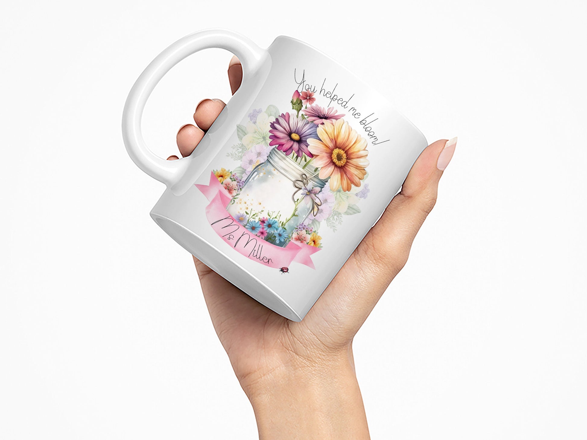White Ceramic mug. A jar vase holding a variety of summer flowers and a pink banner across bottom Text reads You helped me bloom! arched around top of flowers and Name is written within the banner