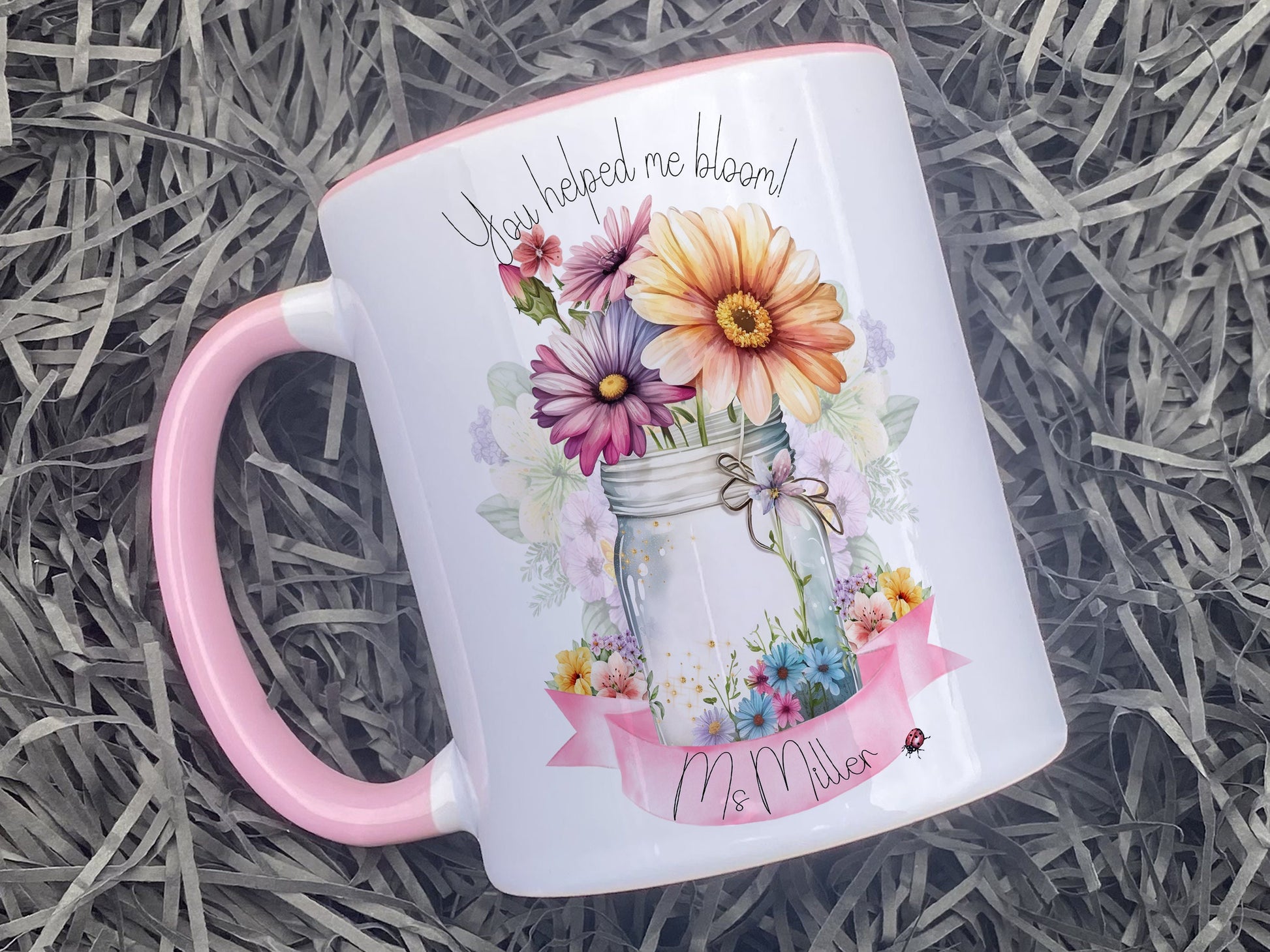 Ceramic mug with pink handle and rim. A jar vase holding a variety of summer flowers and a pink banner across bottom Text reads You helped me bloom! arched around top of flowers and Name is written within the banner