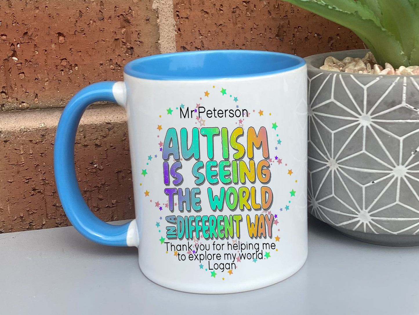 Ceramic mug with blue handle and rim. Bright colourful rainbow wording reading Teachers name and Autism is seeing the world in a differnt way, thank you for helping me to explore my world love name