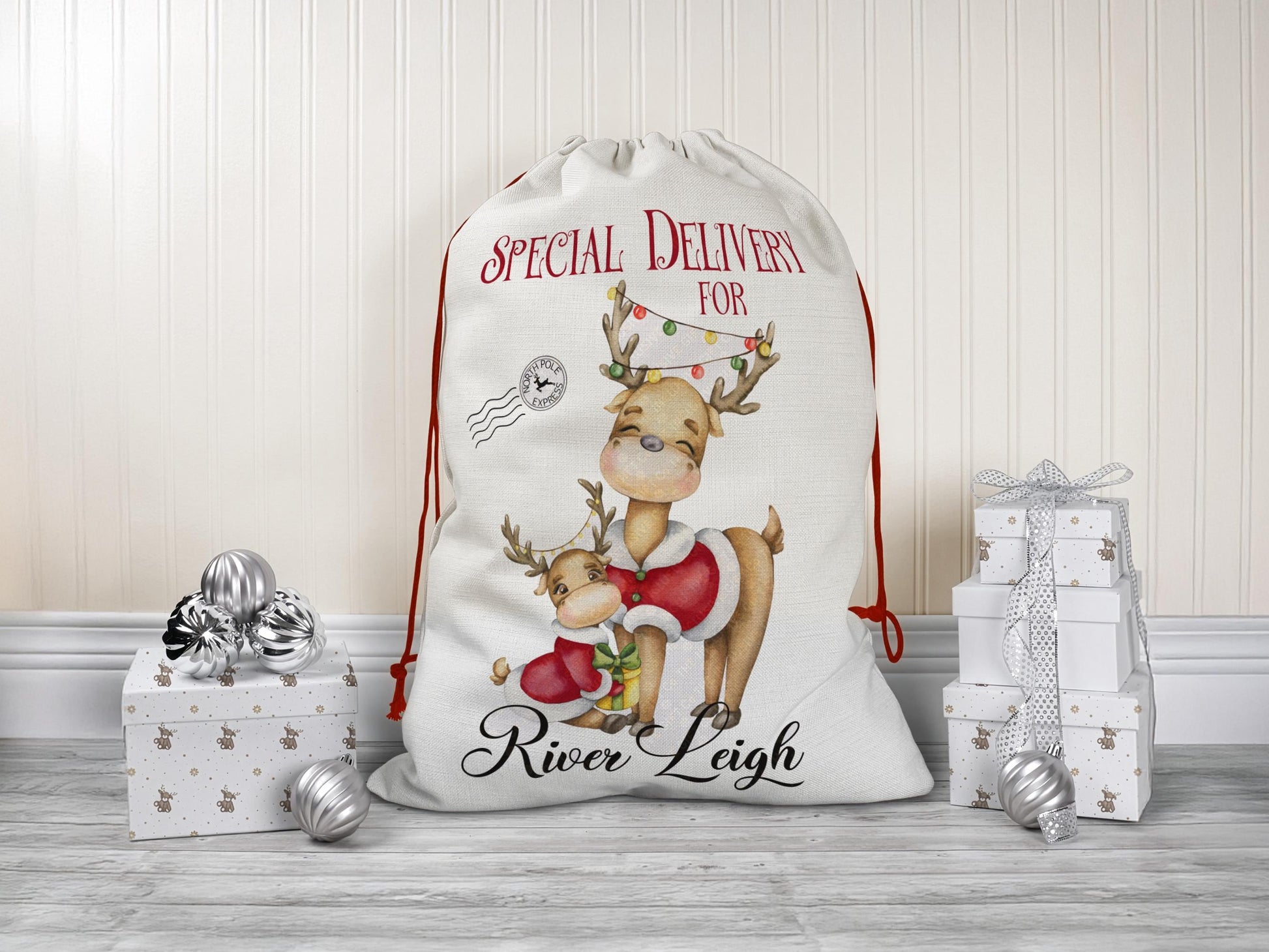 Personalised Christmas Santa sack featuring a cute parent-and-child reindeer illustration with festive lights, "Special Delivery for" text, and custom name "River Leigh" printed at the bottom. The sack has a red drawstring closure and is styled as a gift bag placed among silver ornaments and holiday-wrapped presents.