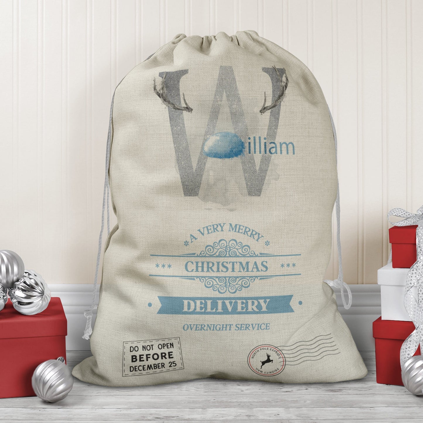 Large natural looking Linen Sack with printed Reindeer antler and Silver Initial letter with blue coloured nose & text Name is written in blue above the words A very merry Christmas Delivery Overnight service and two North pole style delivery stamps
