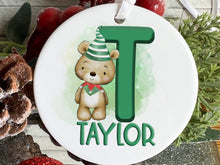Load image into Gallery viewer, White ceramic bauble with Green letter with Cute brown teddy dressed in green and white striped hat and green and red elf collar.  Name written across the bottom in matching Green.
