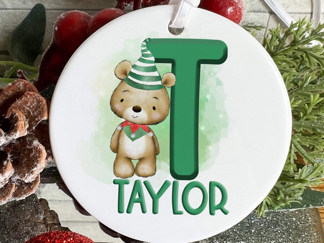 White ceramic bauble with Green letter with Cute brown teddy dressed in green and white striped hat and green and red elf collar.  Name written across the bottom in matching Green.