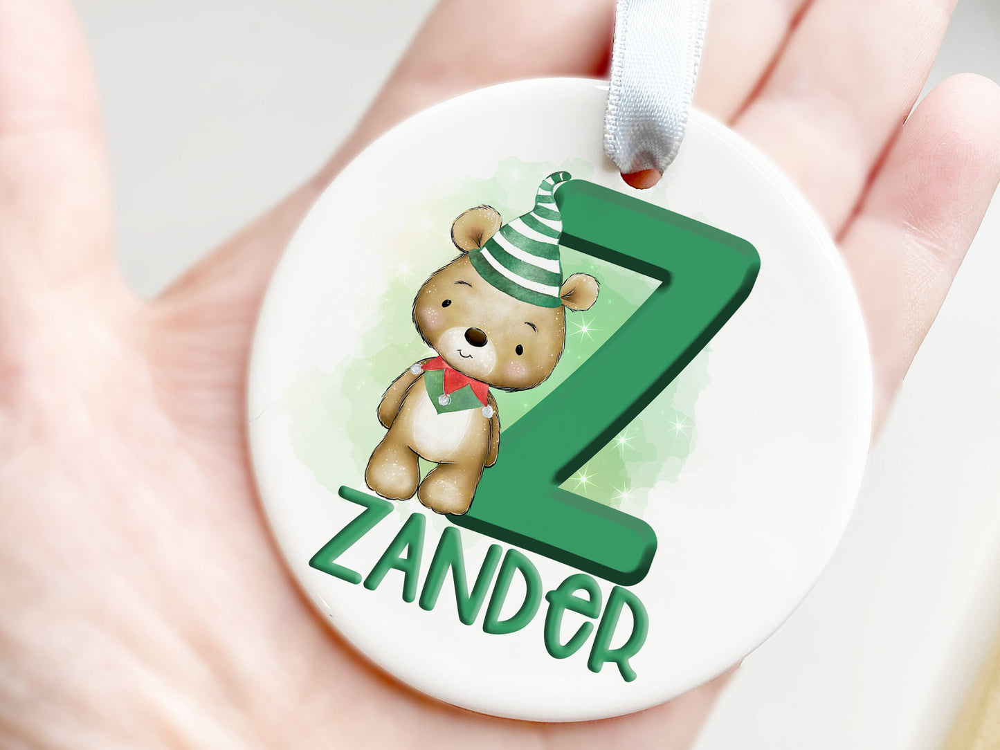 White ceramic bauble with Green letter with Cute brown teddy dressed in green and white striped hat and green and red elf collar.  Name written across the bottom in matching Green.