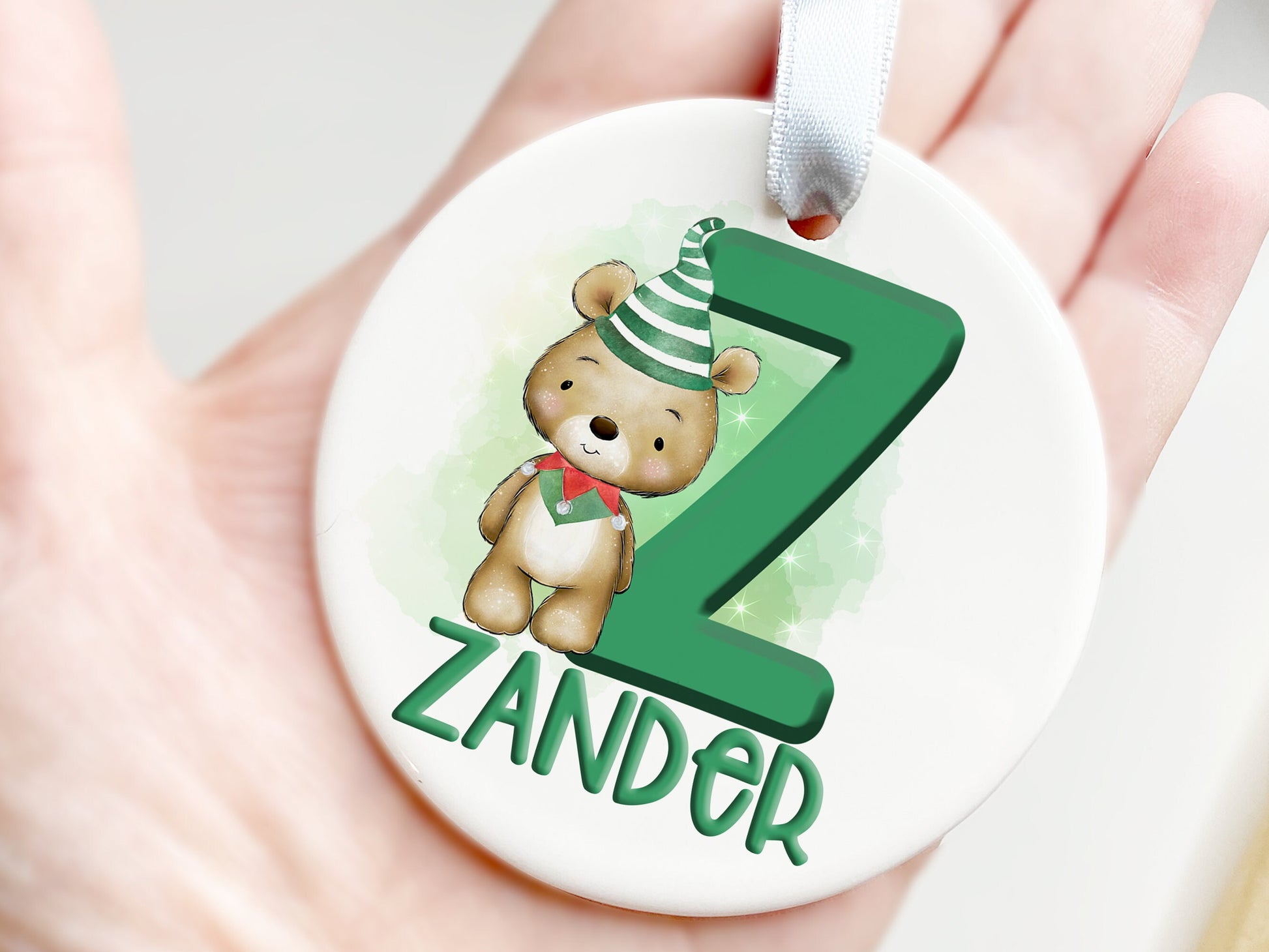 White ceramic bauble with Green letter with Cute brown teddy dressed in green and white striped hat and green and red elf collar.  Name written across the bottom in matching Green.