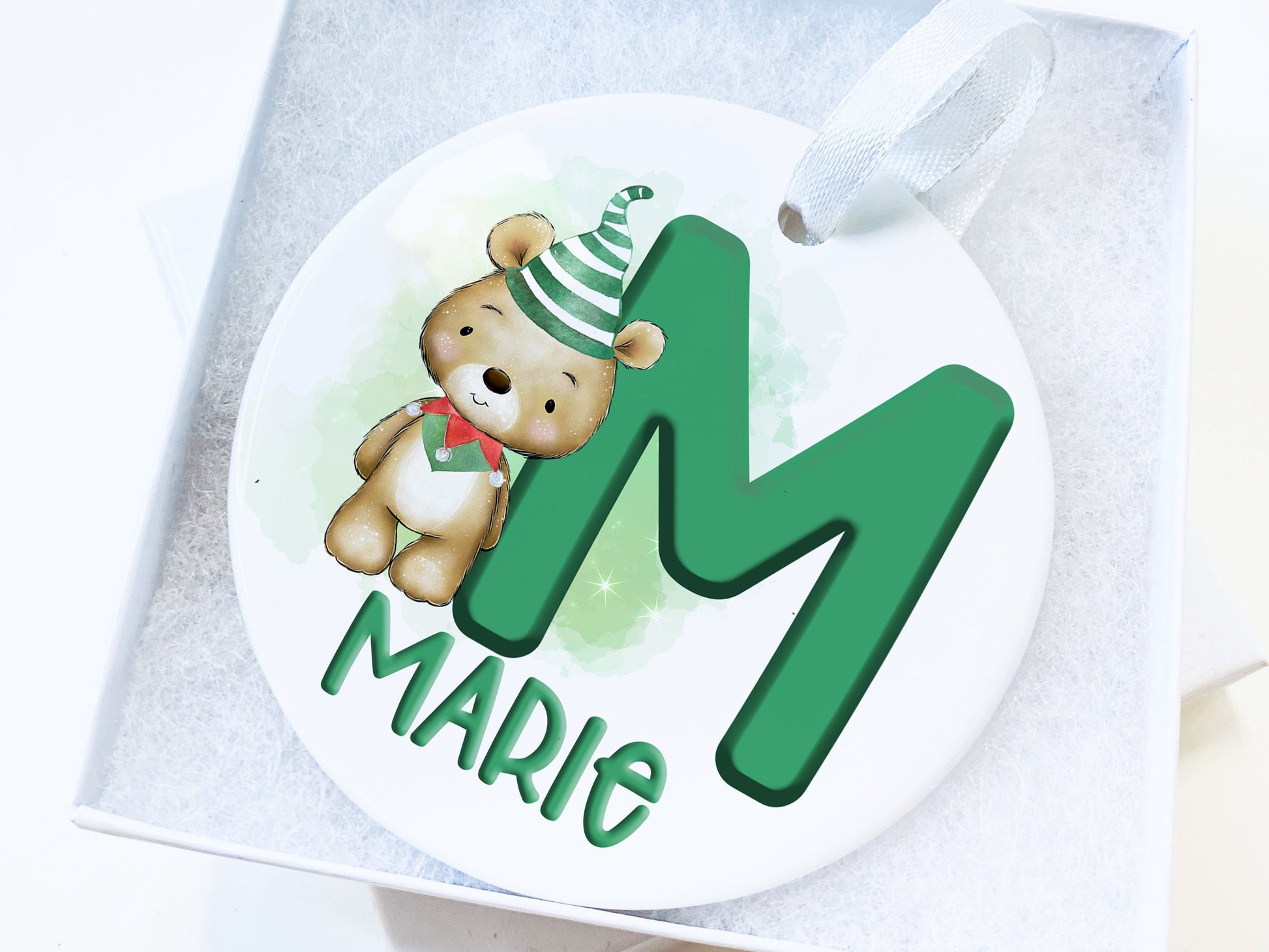 White ceramic bauble with Green letter with Cute brown teddy dressed in green and white striped hat and green and red elf collar.  Name written across the bottom in matching Green. presented in a lovely white gift box