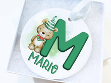 Load image into Gallery viewer, White ceramic bauble with Green letter with Cute brown teddy dressed in green and white striped hat and green and red elf collar.  Name written across the bottom in matching Green. presented in a lovely white gift box
