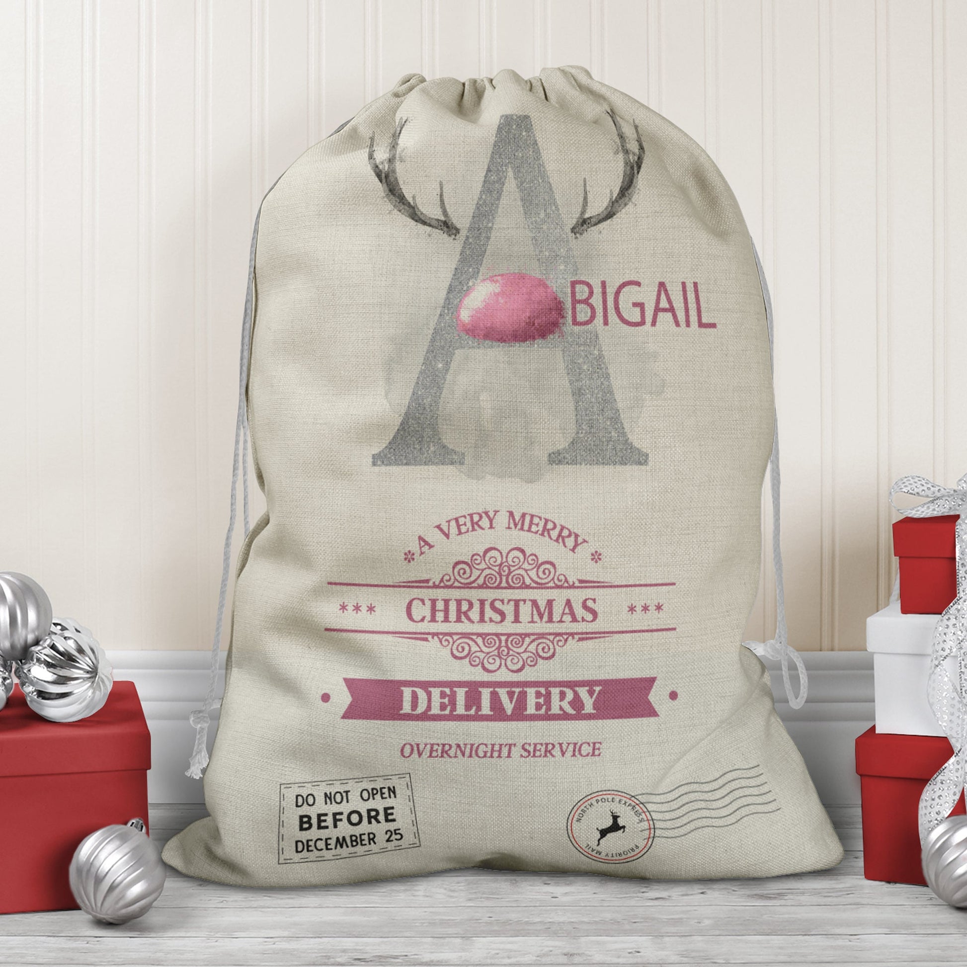 Large natural looking Linen Sack with printed Reindeer antler and Silver Initial letter with pink coloured nose & text Name is written in pink above the words A very merry Christmas Delivery Overnight service and two North pole style delivery stamps
