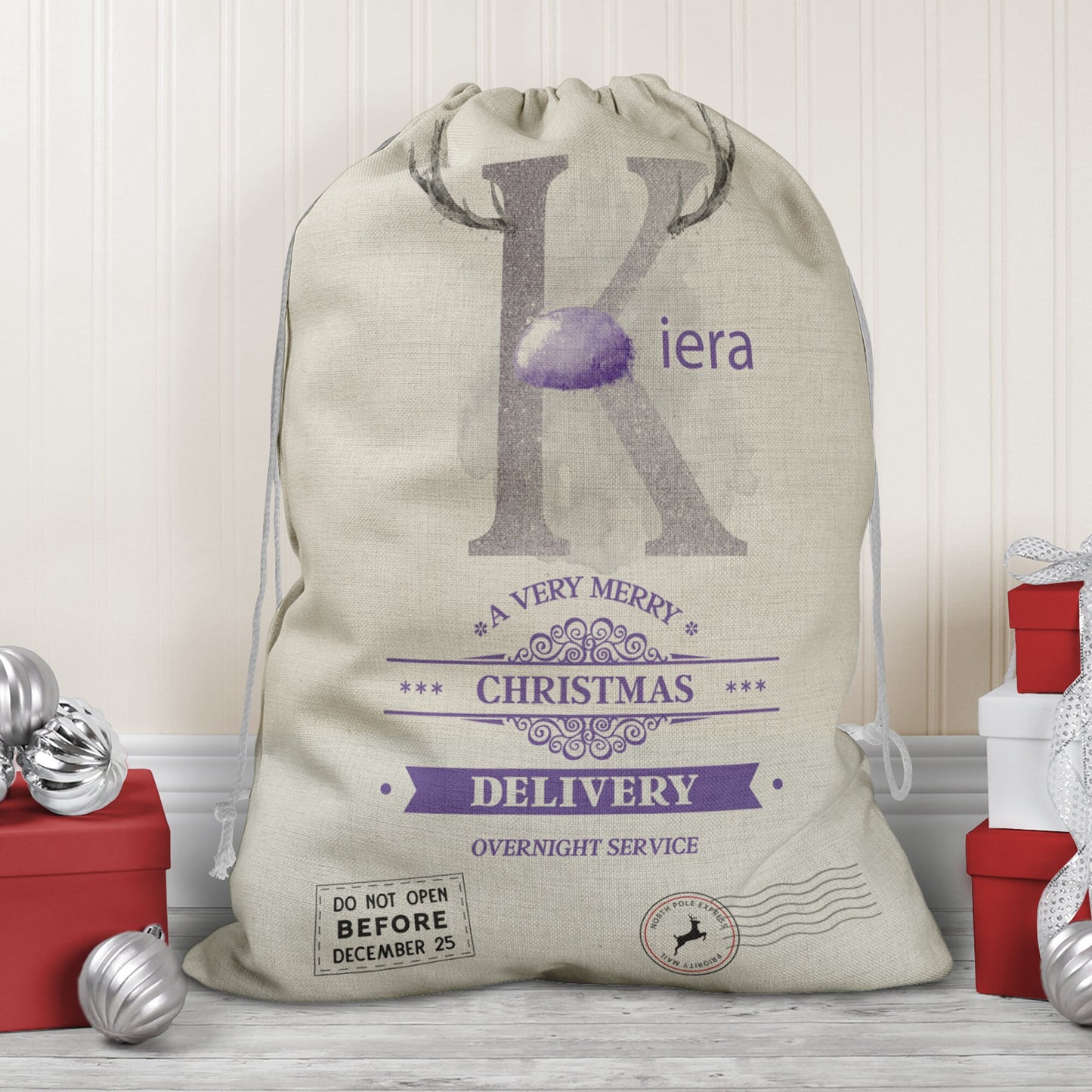 Large natural looking Linen Sack with printed Reindeer antler and Silver Initial letter with lilac coloured nose & text Name is written in lilac above the words A very merry Christmas Delivery Overnight service and two North pole style delivery stamp