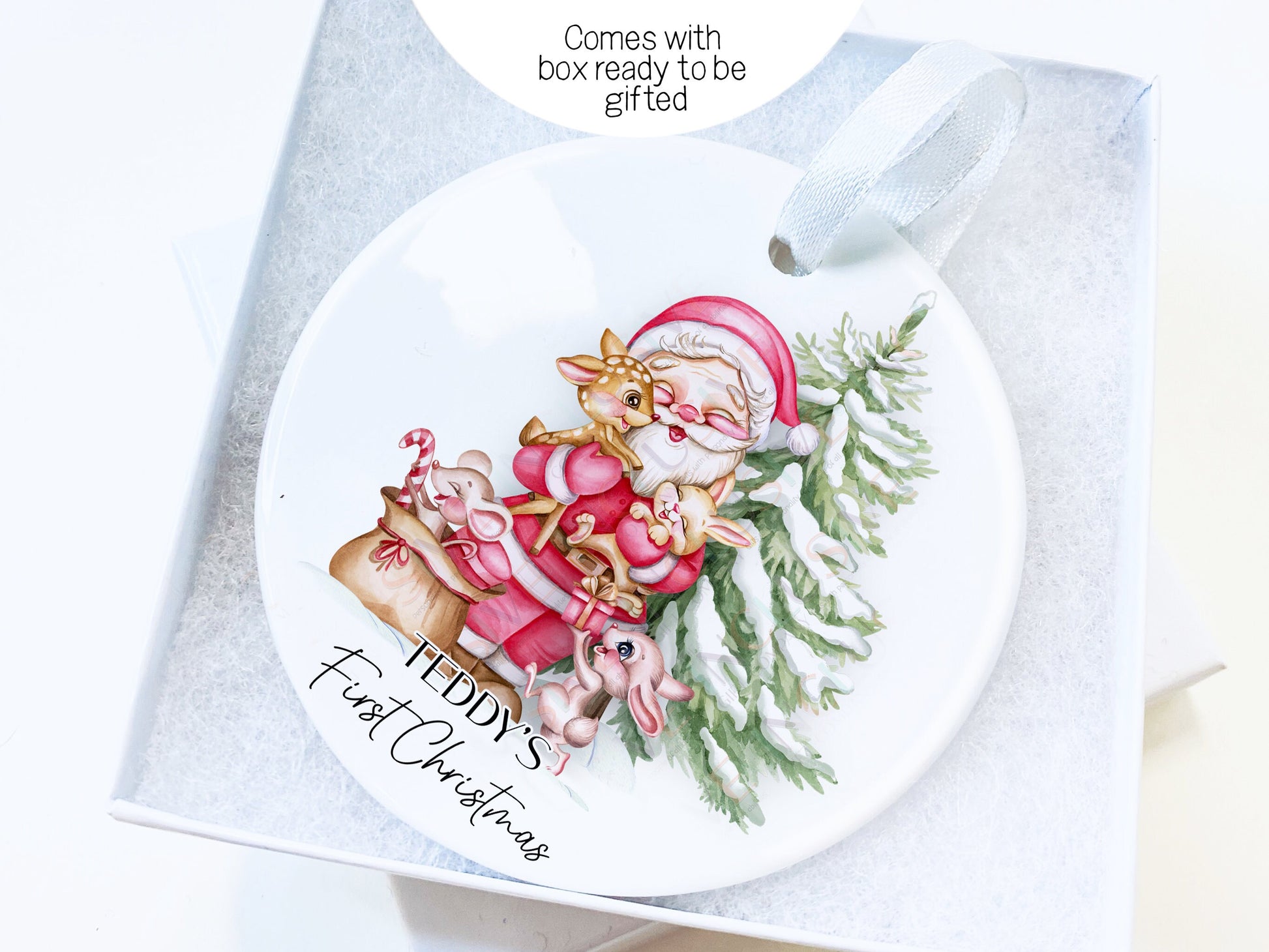 7cm round white ceramic bauble with smiling Santa standing in front of a snowy Pine tree giving a baby deer and baby bunny hugs while another rabbit and mouse play with a sack of presents on snowy ground. Comes with box ready to be gifted