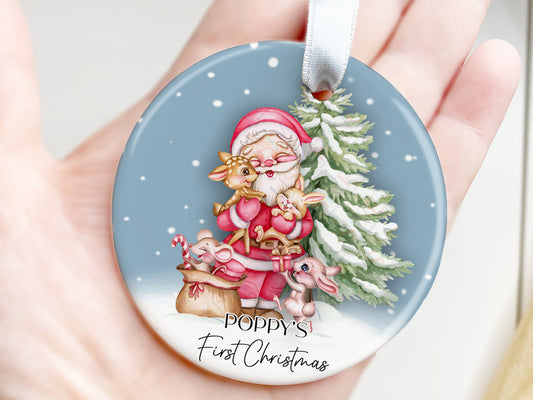 7cm round ceramic bauble with festive snowy blue sky with christmas tree and freshly laid snow. Smiling Santa stands in front of the tree giving a baby deer and baby bunny hugs while another rabbit and mouse play with a sack of presents.