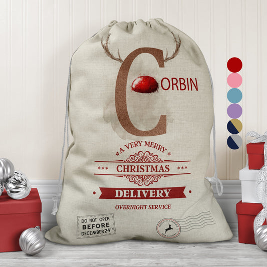 Large natural looking Linen Sack with printed Reindeer antler and Brown Initial letter with red coloured nose, Name is written in red above the words A very merry Christmas Delivery Overnight service and two North pole style delivery stamps