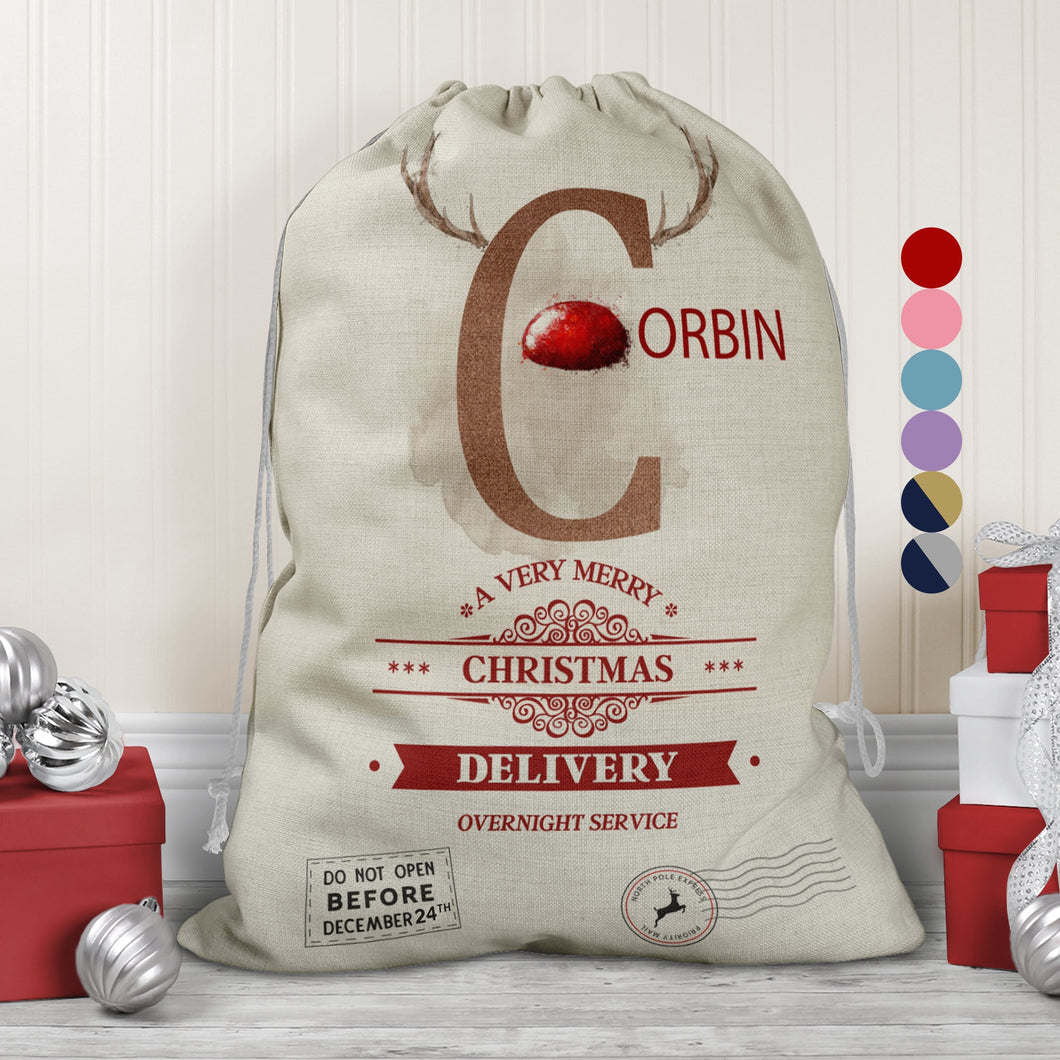 Large natural looking Linen Sack with printed Reindeer antler and Brown Initial letter with red coloured nose, Name is written in red above the words A very merry Christmas Delivery Overnight service and two North pole style delivery stamps