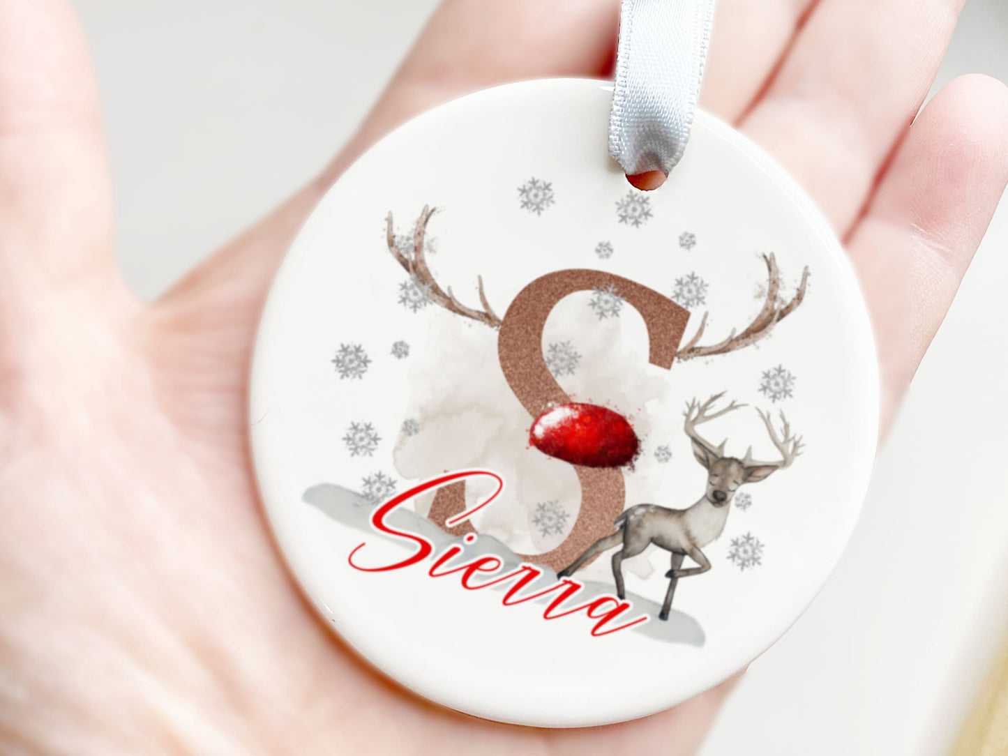 White ceramic bauble with a reindeer standing in the snow with falling snowflakes Personalised with A Bold Initial decorated with a coloured nose and antlers reminiscent of santas magical flying reindeer and Name which matches colour of the nose.