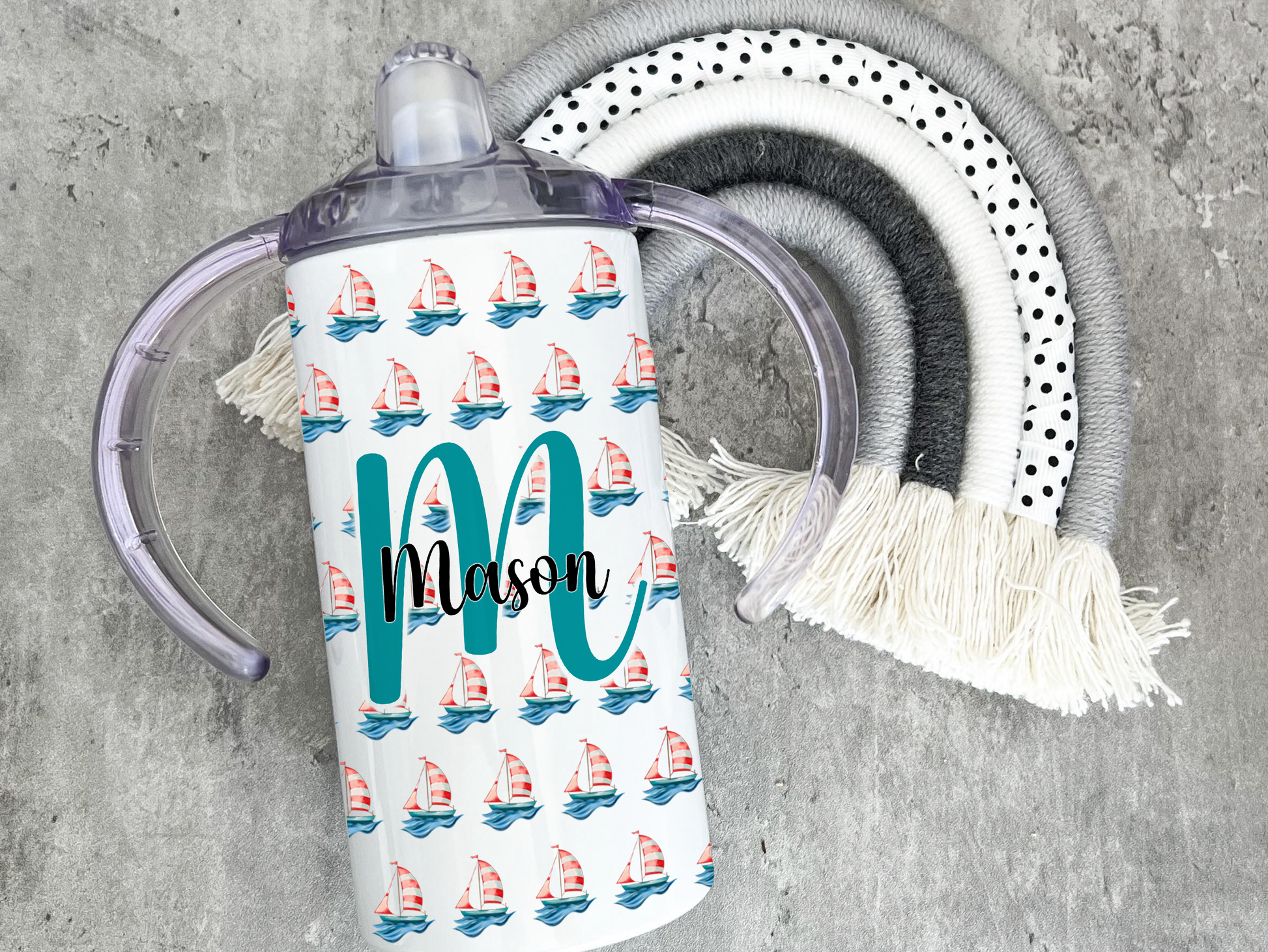 White Adult sippy cup with sailing boat design and inital and name, adaptive cup with Spouted lid for adults with disabilities. Image shows single cup on laying on grey rainbow prop. Design shows sailing boat pattern and Large Initial with name across the middle.